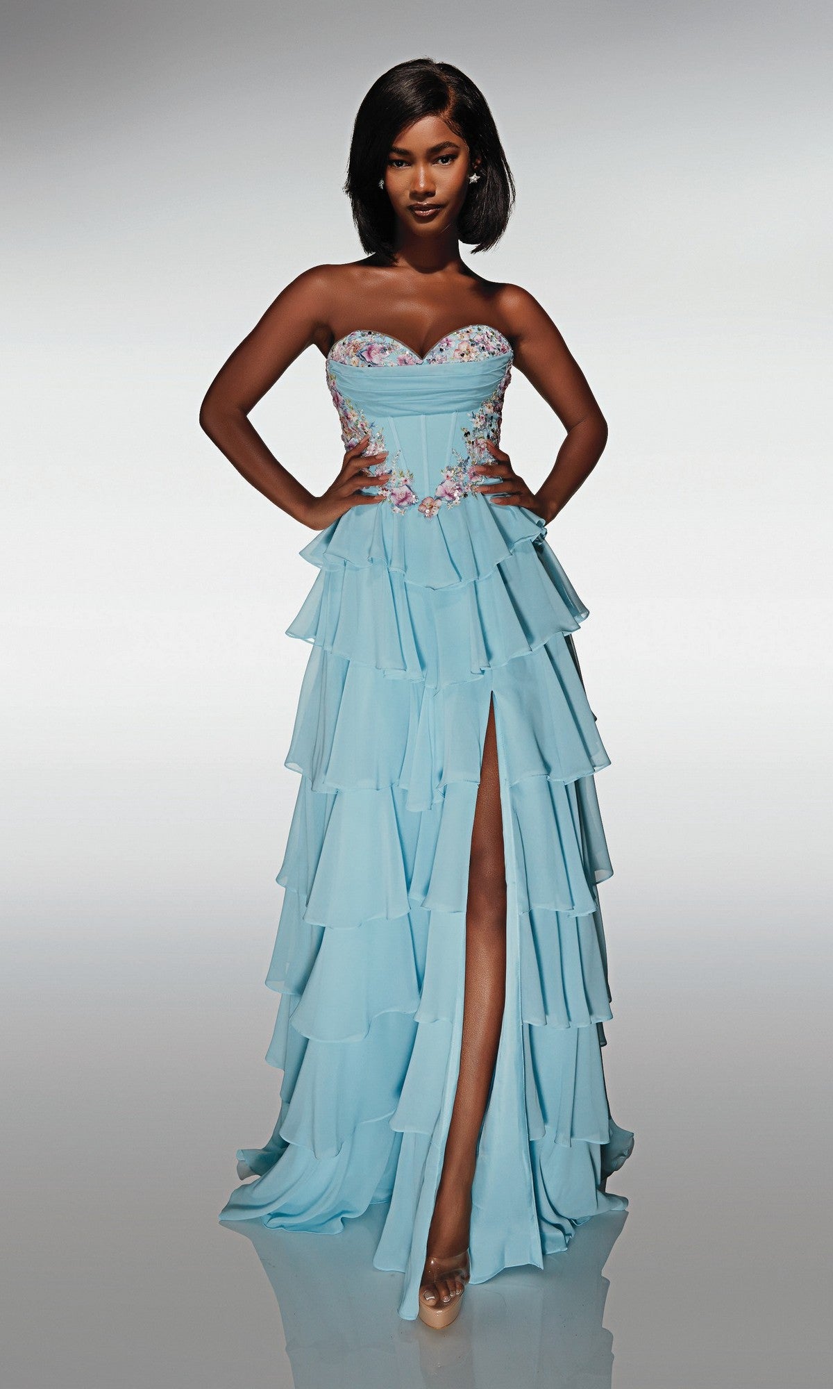 Woman models the Alyce 62013 long prom dress in glacier blue, a strapless chiffon gown with tiered layers, floral appliques on the corset bodice, and a front slit, standing hands on hips against a plain background.